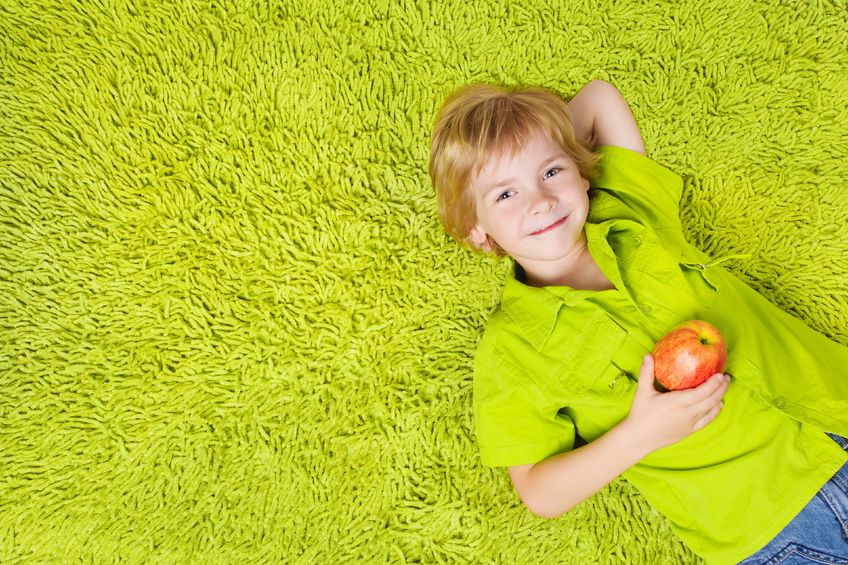 15574677 - child lying on the green carpet background, holding apple. boy smiling and looking at camera